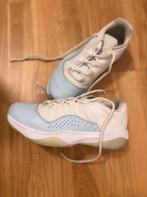 Jordan sneakers i vitt och ljusblått - Snygga sneakers från Jordan i vitt och ljusblått med klassisk låg siluett. Skorna har perforerade detaljer på sidan, vita skosnören och en mjuk, vadderad insida. Ovandelen är i läder och textil, och yttersulan är vit med ljusblå inslag. Perfekta för sport