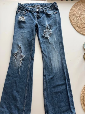 Blå bootcut jeans med hål midjemått 37 - Säljer ett par blå bootcut jeans med slitningar och hål på låren och knäna. Jeansen har klassisk femficksmodell, låg midja och är tillverkade i denim. Perfekta för en avslappnad och trendig look.