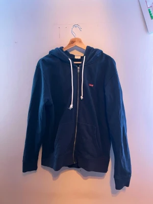 Mörkblå zip hoodie från Levi's - Snygg mörkblå hoodie från Levi's med dragkedja framtill och vita dragsnören i huvan. Klassisk standard fit och två fickor framtill. Liten röd Levi's-logga på bröstet. Perfekt för en avslappnad och cool stil.