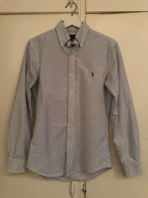 Blårandig skjorta från Ralph Lauren - Klassisk blårandig skjorta från Ralph Lauren med button-down krage och broderad logga på bröstet. Skjortan har långa ärmar, vita knappar och är tillverkad i mjuk bomull. Perfekt för en clean och stilren look.