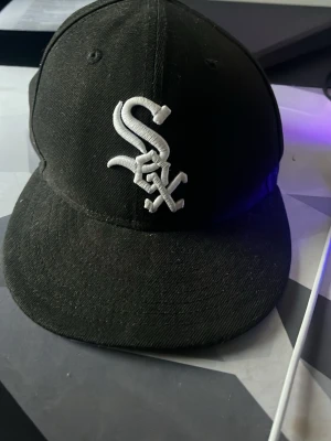 Svart Fitted Sox Keps - Säljer en svart keps från New Era med White Sox broderad logga i vitt framtill. Klassisk rak skärm och justerbar passform. Perfekt för dig som gillar sportig streetstyle och vill ha en stilren accessoar till din outfit.
