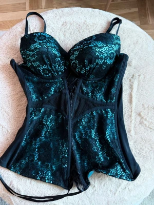 Y2k Svart och turkos spetsbody - Snygg body med vadderade kupor och justerbara axelband. Bodyn är svart med turkos spetsdetaljer och har snörning framtill samt meshpaneler på sidorna. Materialet är en mix av spets och mesh som ger en lyxig känsla. Perfekt för dig som vill ha något extra.