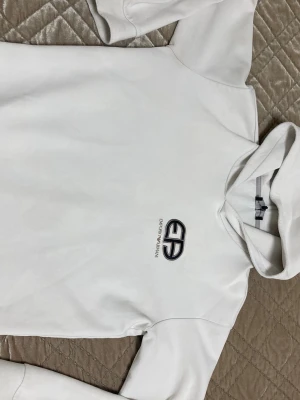 Vit hoodie från Emporio Armani - Snygg vit hoodie från Emporio Armani med svart broderad logga och text på bröstet. Klassisk huva och långärmad modell i mjukt material som känns skönt mot huden. Perfekt för dig som gillar stilrena och exklusiva plagg.