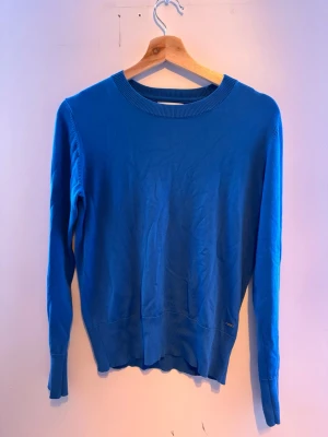 Blå stickad tröja med guldknappar - Blå stickad tröja med rund hals och långa ärmar. Ärmsluten har ribbade muddar och tre snygga guldfärgade knappar som detalj. Tröjan är tunnstickad och har en klassisk, rak passform som funkar till det mesta.