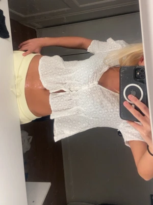 Vit broderad blus med kort ärm - Söt vit blus med broderade hålmönster och korta ärmar. Modellen är croppad och har knappar framtill. Perfekt för en somrig och fräsch look. Materialet känns lätt och luftigt, vilket gör den skön att bära under varma dagar.