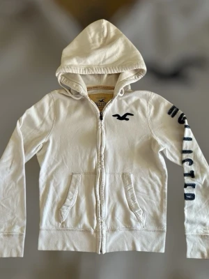  Hollister hoodie - Vit hoodie från Hollister med dragkedja framtill och två fickor. Svart Hollister-logga på bröstet och tryckt text på ena ärmen. Tillverkad i mjuk bomullsblandning och har en klassisk huva för extra komfort.