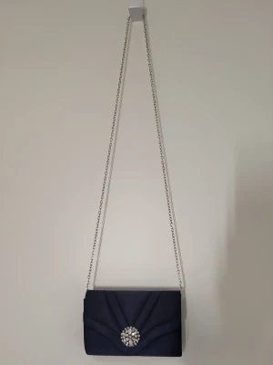 Mörkblå kuvertväska med kedja - Snygg mörkblå kuvertväska med silverfärgad kedja och dekorativt smycke framtill. Väskan har en elegant satinliknande yta och klassisk rektangulär form. Perfekt accessoar för att lyfta din outfit. Har dock tappat en av stenarna.