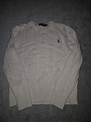 Vit kabelstickad tröja Polo Ralph Lauren - Kabelstickad vit tröja från Polo Ralph Lauren med rund halsringning och diskret broderad logga på bröstet. Klassisk modell med ribbade muddar vid ärmslut och nederkant. Perfekt för en clean och stilren look. Storlek S i fint skick