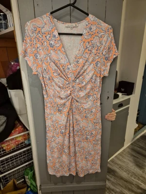 Mönstrad omlottklänning från Indiska - Kortärmad klänning från Flash Woman med paisley-mönster i aprikos, blått och vitt. Klänningen har v-ringning, omlottdetalj framtill och lätt rynkning i midjan för en smickrande passform och ett fint fall. Tillverkad i mjuk och sval viskos, perfekt för varma dagar.