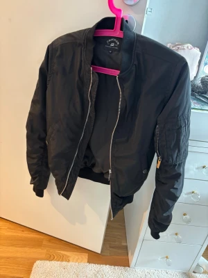 Svart bomberjacka från Crocker - Snygg svart bomberjacka från Crocker med klassisk passform. Jackan har silverfärgad dragkedja, ribbade muddar vid ärmslut och midja samt fickor på sidorna och en ärmficka med dragkedja. Tillverkad i ett slitstarkt syntetmaterial. Storlek xxs