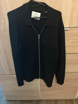 Svart overshirt från Jack & Jones - Stilren svart overshirt från Jack & Jones med dragkedja framtill och två bröstfickor med lock. Klassisk krage och rak passform. Perfekt att bära över en t-shirt för en avslappnad look. Materialet känns mjukt och bekvämt.