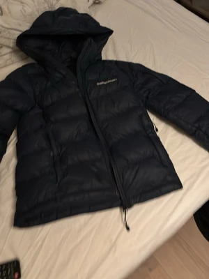 Mörkblå dunjacka Peak Performance - Snygg mörkblå dunjacka från Peak Performance med huva och dragkedja. Jackan har en puffig design och logga broderad på bröstet. Perfekt för kalla vinterdagar och håller dig varm med sitt tjocka dunfoder.