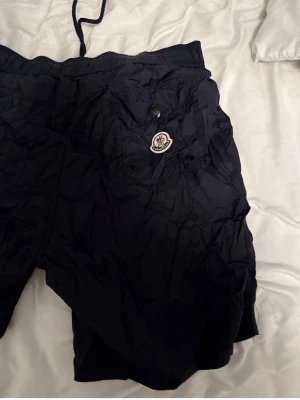 Marinblå badbyxor från Moncler - Säljer ett par marinblå badbyxor från Moncler med klassisk logga på ena benet. De har dragsko i midjan och fickor med knappstängning. Materialet är lätt och snabbtorkande, perfekt för stranden eller poolen.