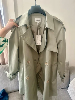 Ljusgrön trenchcoat från Isolde - Stilren ljusgrön trenchcoat från Isolde med klassisk dubbelknäppning, axelklaffar och knytskärp i midjan. Perfekt för dig som vill ha en trendig och tidlös look. Helt ny med prislapp kvar och ej använd! 