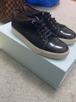 Mörkblåa Lanvin sneakers - Snygga mörblåa sneakers från Lanvin med ovandel i mocka och lackdetaljer på tån. Ett set helt oanvända snören medföljer. Perfekta för dig som gillar en stilren och exklusiv look med en twist av lyx. Perfekta till sommaren. OBS! Storleken passar även 42-43.