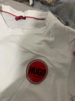 Vit HUGO t-shirt med röd logga - Säljer en vit t-shirt från HUGO med rund hals och korta ärmar. På bröstet finns en stor röd logga med texten 'HUGO WINNING BLEND' och svartvitrutigt mönster runt loggan. Tillverkad i mjuk bomull, perfekt för en clean och stilren look.
