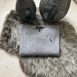 Grå kabelstickad tröja Polo Ralph Lauren - Snygg grå kabelstickad tröja från Polo Ralph Lauren med klassisk rund halsringning och broderad logga på bröstet. Tröjan har långa ärmar och ribbade muddar, perfekt för lager-på-lager under kyliga dagar.