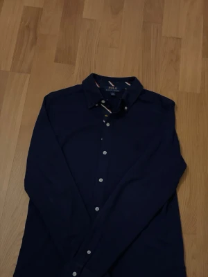 Mörkblå skjorta från Polo Ralph Lauren - Snygg mörkblå tunn skjorta från Polo Ralph Lauren med klassisk krage och vita knappar. Skjortan har diskreta detaljer i kragen med röd och vit rand samt en liten gul broderad logga. Perfekt till jeans eller chinos.