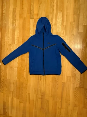 Nike Tech Blå  - Snygg blå hoodie från Nike med hel dragkedja framtill och svarta detaljer. Tröjan har huva, två sidofickor och en extra ficka på ena ärmen. Tillverkad i mjukt material som passar perfekt till vardagslooken. Nike-logga på bröstet för en sportig vibe.