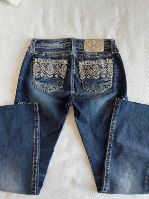Miss Me blåa jeans med mönstrade fickor - Säljer ett par blå Miss Me jeans med snygga slitningar och broderade bakfickor med stenar och mönster. Jeansen har bootcut-modell. Materialet är klassisk denim med stretch för skön passform. Perfekt för dig som gillar detaljer och en unik stil. 💕Innerbensmått 74cm Midjemått tvärs över 37cm Midjehöjd 22cm Benvidd nertill 20,5cm 