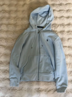 Ljusblå hoodie från Polo Ralph Lauren - Snygg ljusblå hoodie från Polo Ralph Lauren med dragkedja, huva med dragsko och klassisk logga broderad på bröstet. Mjuk bomullskvalitet och känguruficka framtill. Perfekt för en chill och stilren look. I helt nyskick har använts ett par gånger bara! 