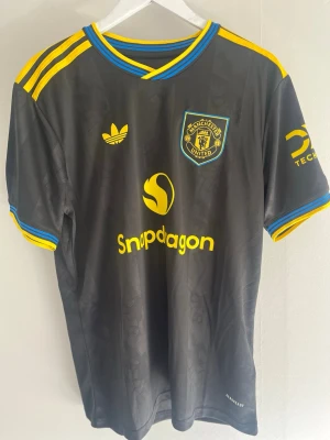 Manchester United Cunha 10 Adidas tröja - Snygg svart Manchester United matchtröja med gula och blå detaljer. Tröjan har tryck med Snapdragon och Tech, samt spelarnamn Cunha och nummer 10 på ryggen. Tillverkad i lätt och ventilerande material, perfekt för fotbollsträning eller match.