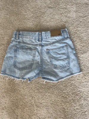 Ljusblå denimshorts från 157 - Säljer ett par ljusblå denimshorts från 157 med råa kanter och slitna detaljer framtill. Shortsen har fem fickor, bälteshällor och knappgylf. Perfekta för sommaren och ger en avslappnad look.