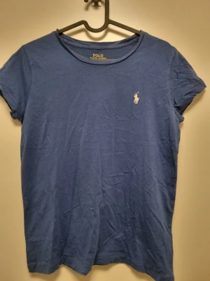 Blå t-shirt från Polo Ralph Lauren - Snygg blå t-shirt från Polo Ralph Lauren med klassisk passform och korta ärmar. T-shirten har en liten broderad logga i vitt på bröstet och är tillverkad i mjuk bomull. Perfekt till jeans eller shorts för en avslappnad stil. Storlek S. 