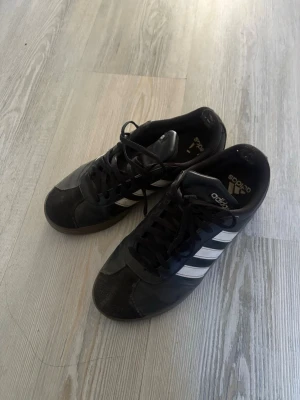Adidas Samba svarta sneakers - Klassiska Adidas Samba sneakers i svart med vita ränder på sidorna och svart snörning. Skorna har låg profil och är tillverkade i en mix av läder och mocka med gummisula. Perfekta för en sportig och stilren look.