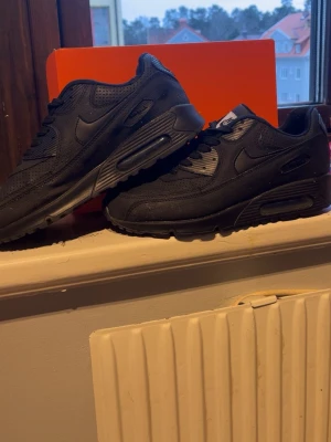 Nike Air Max 90 helsvarta sneakers - Snygga helsvarta Nike Air Max 90 sneakers med klassisk siluett. Skorna har ovandel i mesh och läder, synlig Air Max-dämpning i sulan och diskreta Nike-loggor på sidorna. Perfekta för dig som gillar en clean och sportig look.