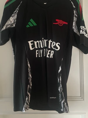 Arsenal 24/25 Gabriel 6 - Snygg svart Arsenal tröja med gröna och röda detaljer vid kragen. På ryggen så står det Gabrie loch nummer 6 i vitt, samt Premier League-logga. Storlek small, perfekt för fotboll eller träning.