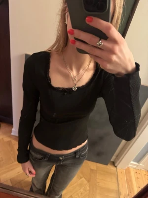 Svart ribbad topp med spets och rosett - Säljer en svart långärmad topp med ribbad struktur. Toppen har en söt liten rosett och spetsdetaljer vid den fyrkantiga ringningen. Sitter tight och är croppad, perfekt till jeans eller kjol.