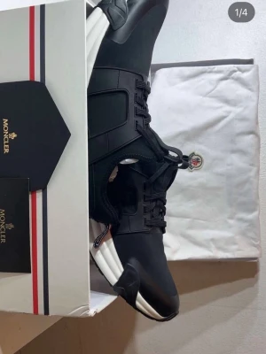 Svarta Moncler sneakers med vit sula - Stilrena svarta sneakers från Moncler med chunky vit sula och svarta snören. Skorna har en modern design med detaljer i gummi och textil, samt Moncler-logga på hälen. Perfekta för dig som gillar exklusiva och trendiga sneakers. 42-43