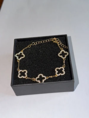 Guldfärgat armband med pärldetaljer - Snyggt guldfärgat armband med fem fyrklöverformade länkar dekorerade med små vita pärlor. Kedjan är tunn och justerbar med karbinlås. Perfekt accessoar för att lyfta din outfit med en elegant touch.