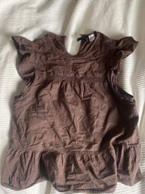 Brun volangblus med spetsdetaljer - Söt brun blus med volangärmar och spetsdetaljer över bröstet. Blusen har en lös passform och volang nertill för en trendig look. Perfekt att styla med jeans eller kjol för en chill vibe.