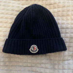Mörkblå ribbstickad Moncler mössa - Säljer en mörkblå ribbstickad mössa från Moncler med uppvikt kant och klassisk Moncler-logga framtill. Mössan är tillverkad i mjukt material och har en stilren, enkel design som passar perfekt till kalla dagar.