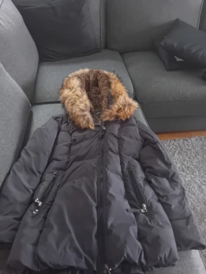 Svart dunjacka med fuskpäls från Hollies - Snygg svart dunjacka från Hollies med stor huva kantad av brun fuskpäls. Jackan har dragkedja framtill, två fickor med dragkedja och quiltad design. Perfekt för kalla vinterdagar och riktigt varm tack vare fodret och den rejäla huvan.