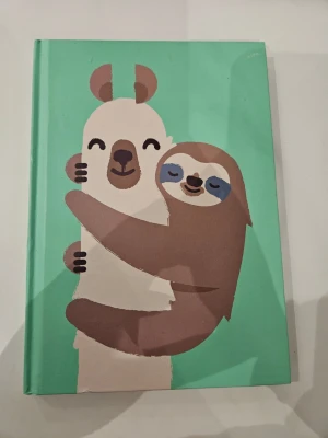 Grön anteckningsbok med djurmotiv - Söt anteckningsbok med hårt grönt omslag och illustrationer av en björn och en sengångare som kramas på framsidan och en glad sengångare på baksidan. Perfekt för anteckningar, dagbok eller skisser. Omslaget är gjort av kartong med tryckt motiv.