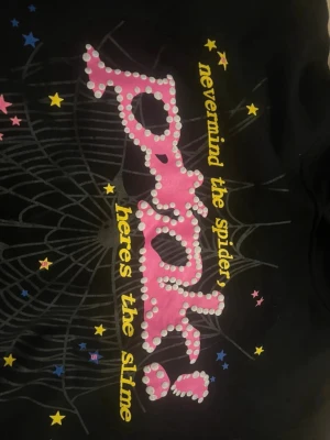 Svart hoodie SP5R med tryck - Cool svart hoodie från SP5R med färgglatt tryck framtill. Texten 'nevermind the spider, heres the slime' i gult och rosa, samt spindelnätsmönster och stjärnor i flera färger. Hoodien har huva och är perfekt för dig som gillar unik streetwear.