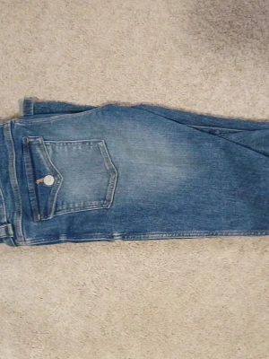 Blå fler jeans med fickdetalj - Säljer ett par klassiska blå jeans som är fler i benen och snygg fickdetalj bak. Jeansen har en ljus tvätt och är tillverkade i denim. Perfekta för en avslappnad och trendig look.