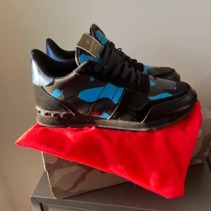 Valentino Garavani Rockrunner sneakers - Snygga Valentino Garavani Rockrunner sneakers i svart och blått med coolt kamouflagemönster. Skorna har svarta snören, detaljer i mocka och skinn samt ikoniska nitar på hälen. Perfekta för dig som vill sticka ut med en lyxig streetstyle. Låda och dust bags medföljer ej!