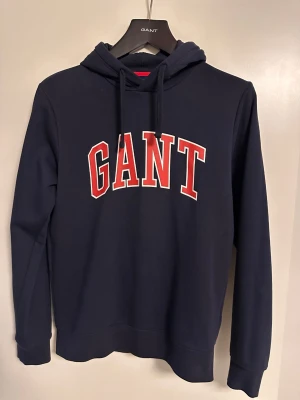 Mörkblå hoodie från GANT med tryck - Snygg mörkblå hoodie från GANT med stor röd och vit logga på bröstet. Klassisk huva med dragsko och mjukt material som känns skönt mot huden. Perfekt för en avslappnad och sportig stil.