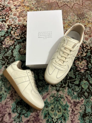 Maison Margiela vita sneakers - Snygga vita sneakers från Maison Margiela i mjukt läder med klassisk gummisula. Modellen har lågt skaft, ton-i-ton sömmar och stilrena detaljer. Perfekta för dig som gillar minimalistisk och clean stil.