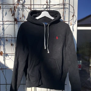 Svart hoodie från Polo Ralph Lauren - Svart hoodie från Polo Ralph Lauren med klassisk röd logga broderad på bröstet. Hoodien har vit dragsko i huvan och en stor magficka framtill. Tillverkad i mjukt bomullsmaterial och har ribbade muddar vid ärmslut och nederkant. Storlek M men passar även S, pris kan diskuteras vid snabbaffär!