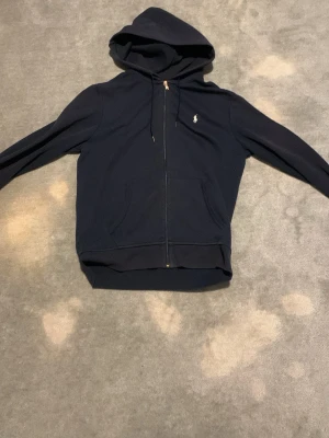 Mörkblå hoodie från Ralph Lauren - Snygg mörkblå hoodie med dragkedja från Ralph Lauren. Klassisk design med huva, två fickor framtill och den ikoniska vita Polo-loggan broderad på bröstet. Perfekt för en avslappnad och stilren look.