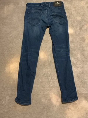 Blå slim fit jeans från Replay - Snygga blå slim fit jeans från Replay med klassisk femficksdesign och diskreta slitningar. Jeansen har normal midja och raka ben, tillverkade i mjuk denim med lite stretch för extra komfort. Perfekta för en avslappnad och trendig look.