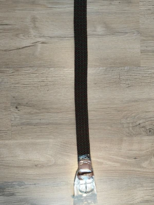 Brunt flätat bälte med läderdetaljer - Snyggt flätat bälte i mörkbrunt med läderdetaljer vid spännet. Bältet har ett klassiskt metallspänne och passar perfekt till jeans eller chinos. Stilen är enkel men trendig och funkar till många olika outfits.