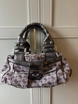 Lila mönstrad handväska från Guess - Cool handväska från Guess i lila och grått/beige tyg med GG-logga över hela väskan. Handtag och detaljer i svart, och reptilmönster. Väskan har en dekorativ spänndetalj framtill och silverfärgade metalldetaljer.