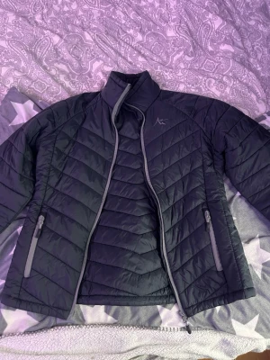 Svart pufferjacka från Quechua - Snygg svart pufferjacka från Quechua med grå dragkedjor och logga på bröstet. Jackan har hög krage, två sidofickor med dragkedja och quiltad design. Perfekt för kalla dagar och enkel att matcha med olika outfits.