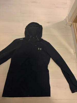 Svart vindjacka från Under Armour - Snygg svart vindjacka från Under Armour med huva och dragsko. Jackan har dragkedja framtill, en bröstficka med dragkedja och logga på vänster sida. Perfekt för blåsiga dagar och sportiga outfits.